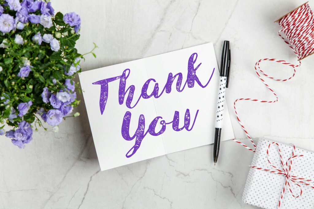 A stylish thank you card paired with purple flowers, a gift box, and pen on a marble surface.